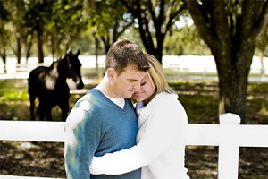 Couple in loving embrace, standing in front of white fence, horse in background