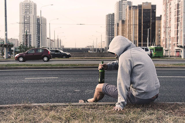 Man in hoodie sitting on a city street curb drinking a beer