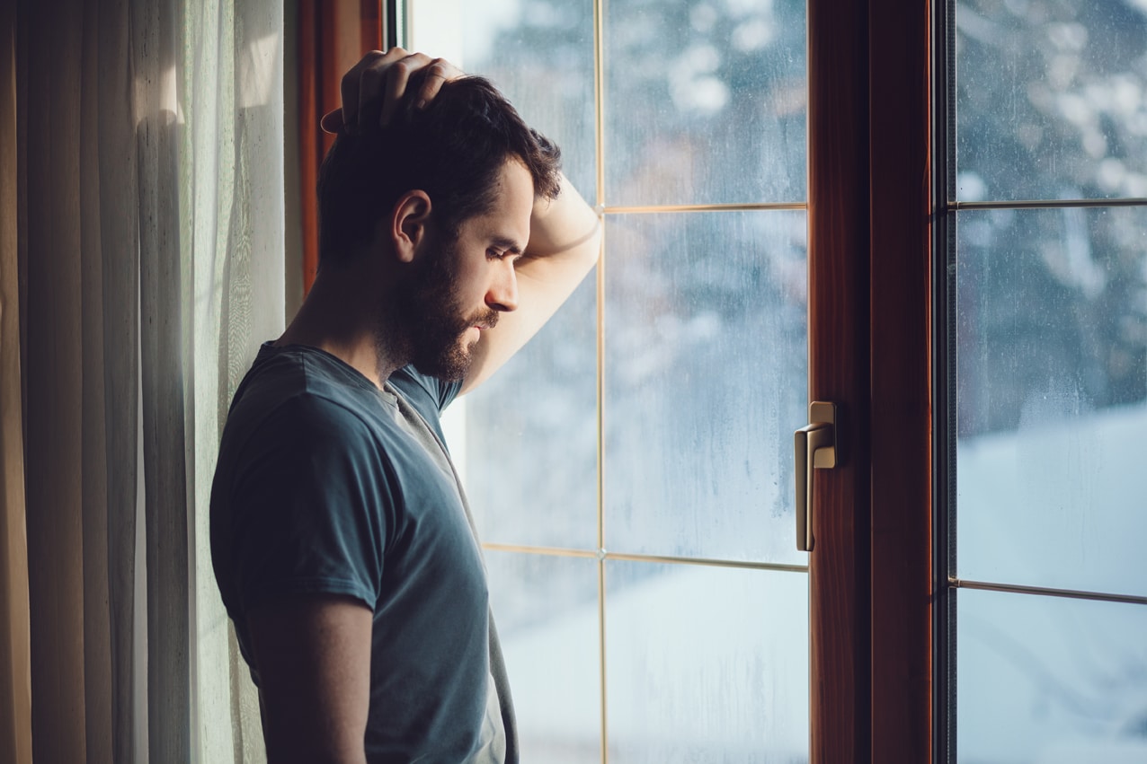 A young man with a beard leaning against a window with a hand on his head and a sad look on his face.