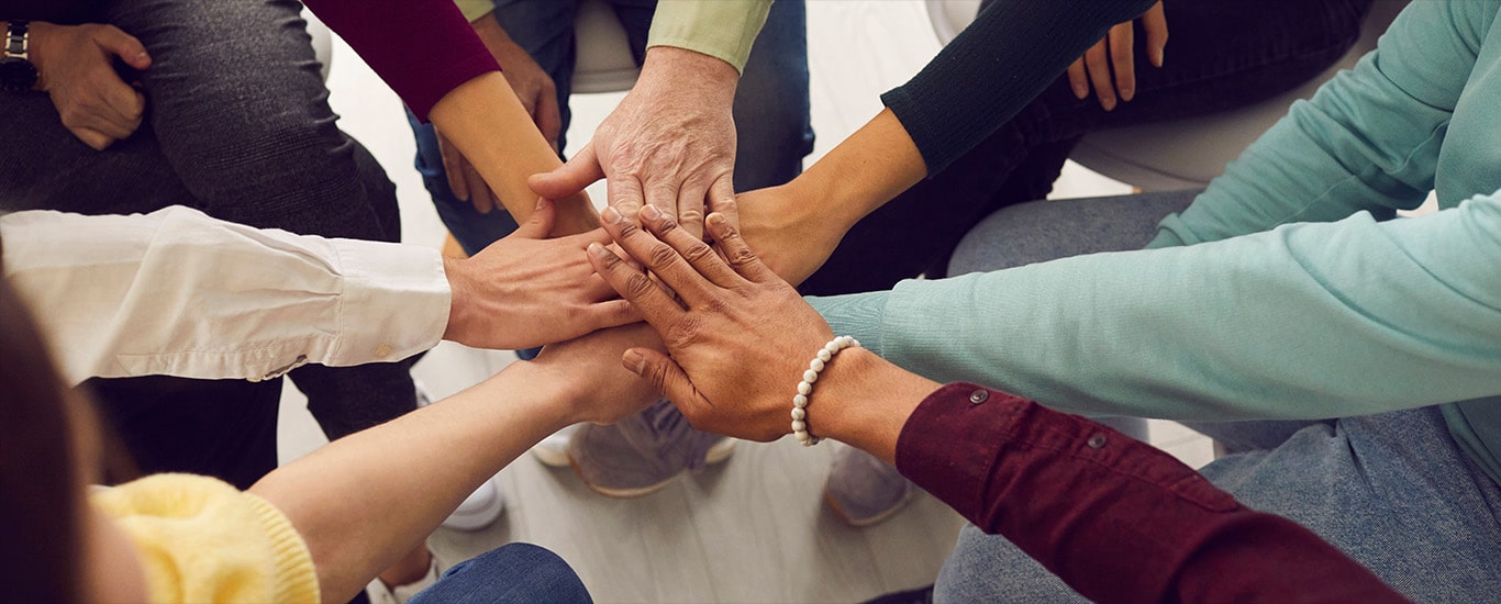 Several people sit in circle and put their hands together in a hand-stack during a group therapy session.