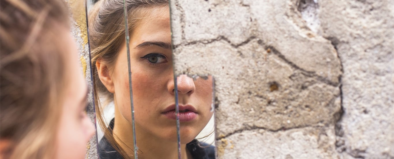 A pensive young woman stares at her reflection in a broken mirror.