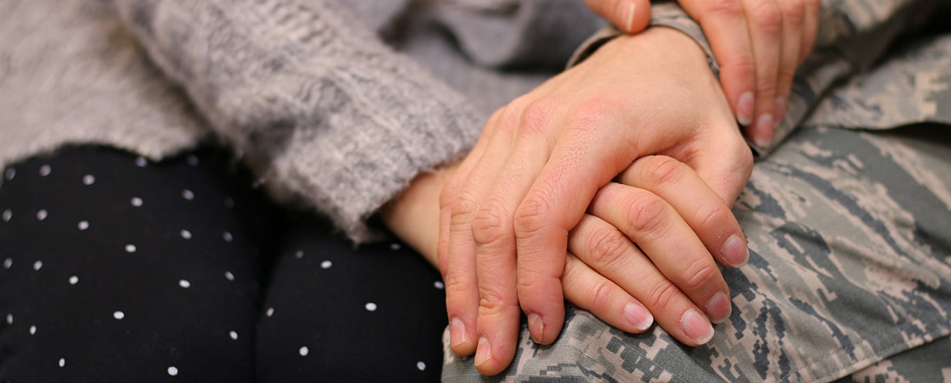 A young man and woman hold hands to comfort one another. The man wears army fatigues