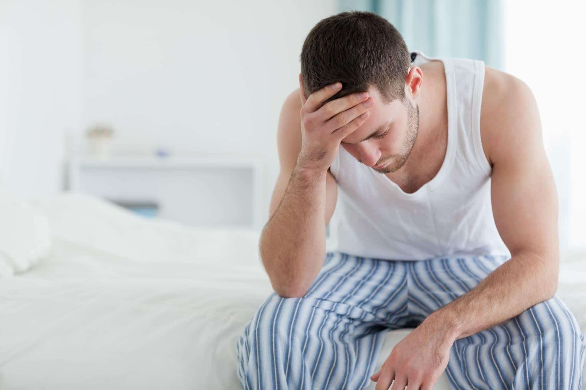 A sick man in a white tank top and striped pajama pants sits on the corner of his bed, holding his forehead in pain.