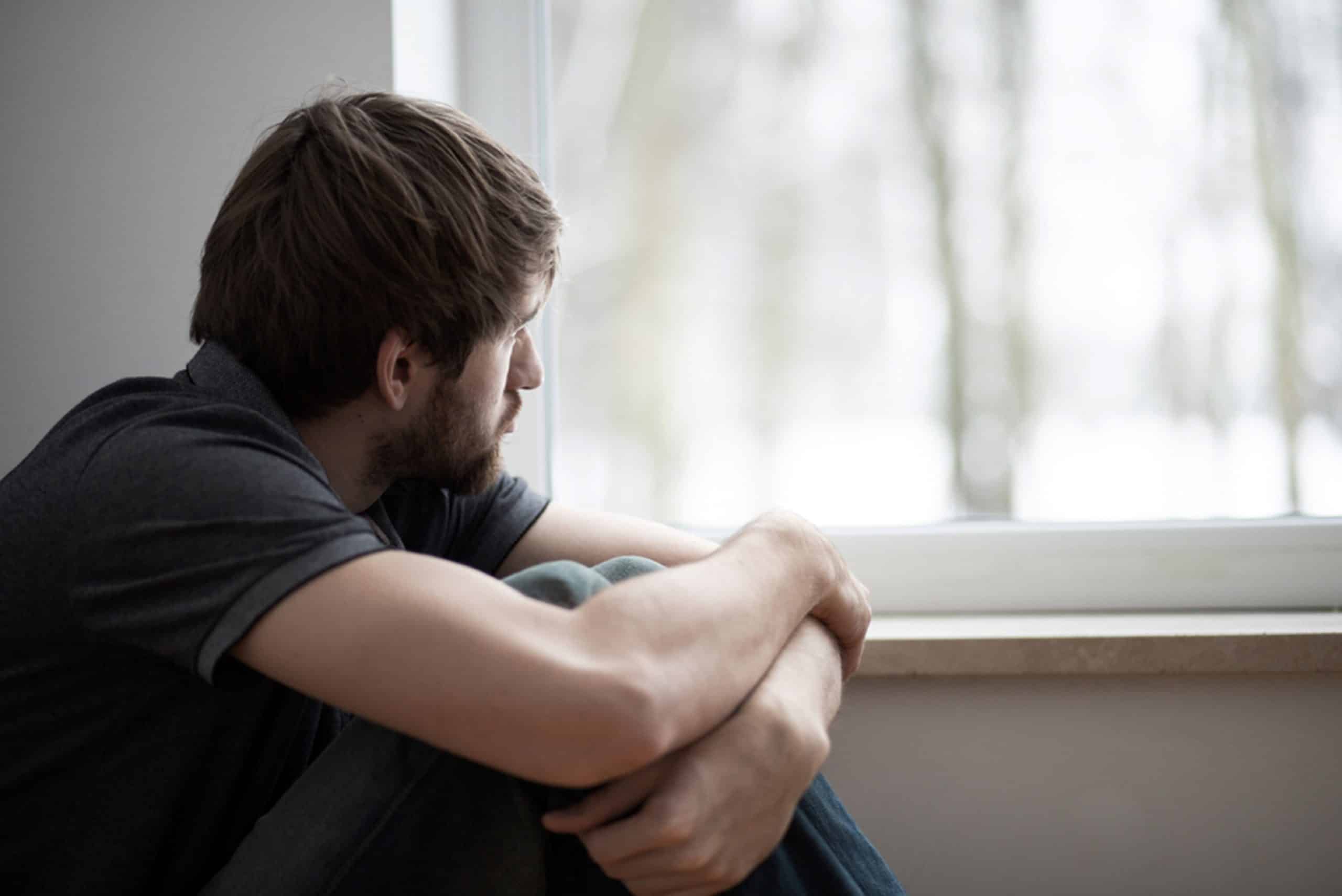 Sad young man sitting on the floor with his arms around his knees looking out the window.