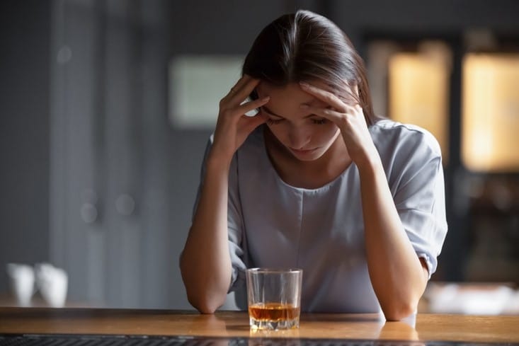 A young woman is sitting at a bar with an alcoholic beverage in front of her holding her hands to her forehead with a sad demeanor on her face.