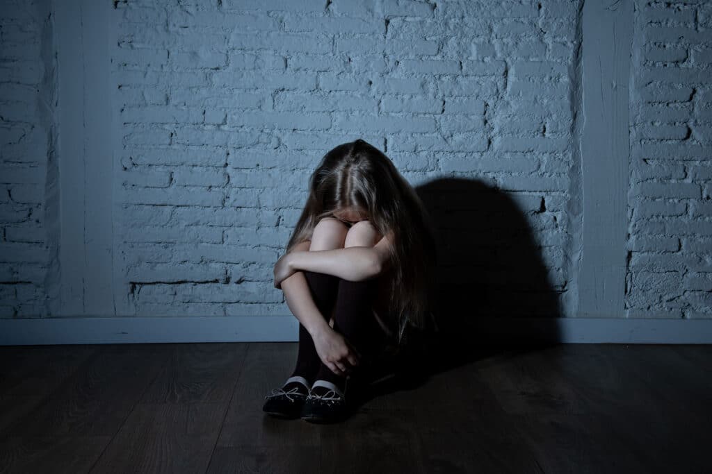 A young girl sits in a dark room against a white wall with her arms around her bent knees and her face hidden in her forearms.