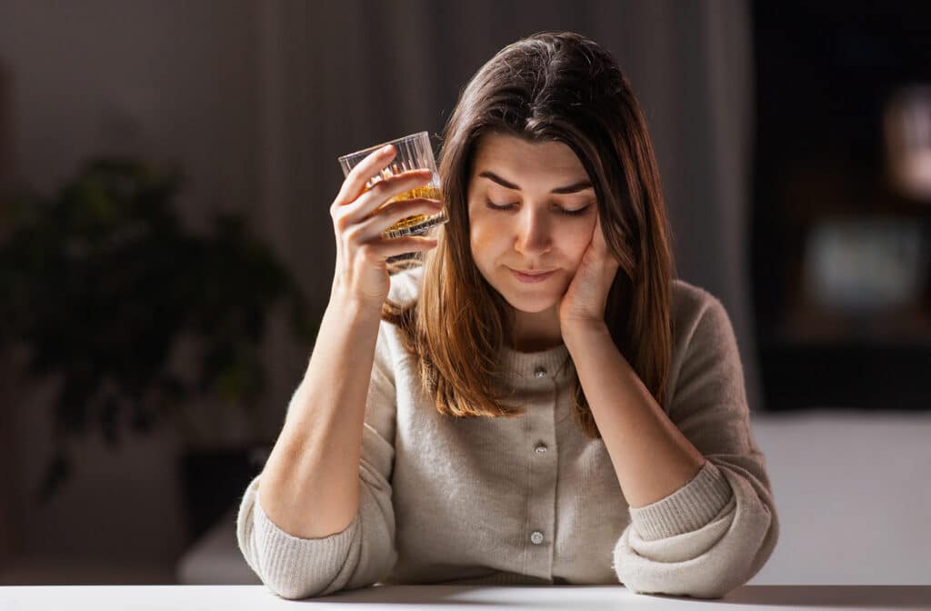 A woman sits at a table with her arms up, one hand holding a cup of whiskey and the other on her cheek